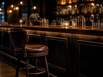 Dimly lit bar with a dark wooden counter and a brown leather barstool in the foreground, warm pendant lights and bottle shelves in the background.
