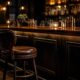 Dimly lit bar with a dark wooden counter and a brown leather barstool in the foreground, warm pendant lights and bottle shelves in the background.